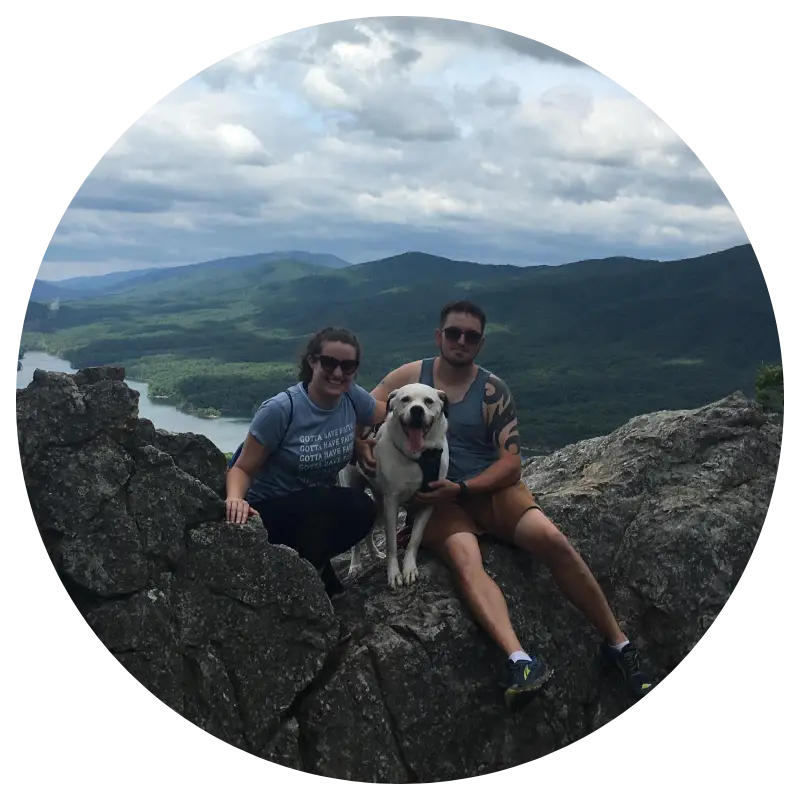 A couple sits on a rocky ledge beside a smiling dog, with scenic mountains and a river in the background under a cloudy sky.